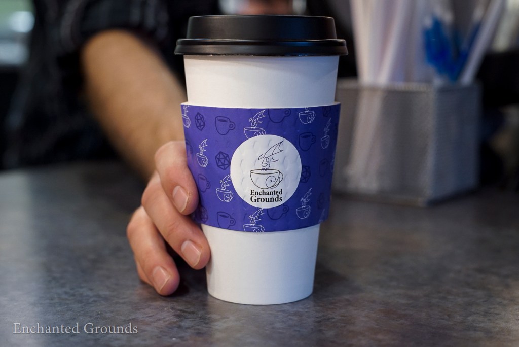 A photo of a paper coffee cup, seemingly pushed forward by a barista's hand. The cup sleeve has an Enchanted Grounds logo on it, with a blue background and repeating pattern of cups, dice, and the EG logo.